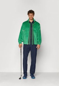 Helles grünes Cordjackett mit Reißverschluss vorne, kombiniert mit einem marineblauen Polo mit grünen Streifen und dunkler Hose, hält einen Golfschläger.