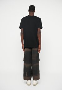 Homme portant un t-shirt noir, un pantalon cargo large marron et noir, et des baskets blanches, se tenant de dos contre un fond blanc.