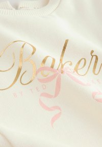 Krémová fleece tkanina má zlaté písmená s nápisom "Pekár" a dizajn s ružovou stuhou. Tkanina má mäkkú štruktúru a ribbovaný výstrih.