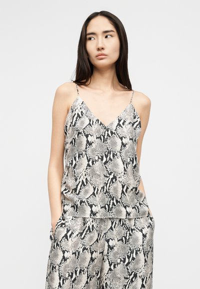 Γυναίκα με μακριά σκούρα μαλλιά, φοράει αμάνικο τοπ και παντελόνι με μοτίβο μαύρης και λευκής δερματίνης φιδιού, με τα χέρια στις τσέπες, κοιτάζει αριστερά.