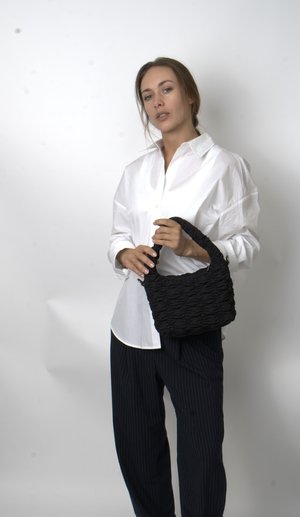 Frau in weißem Oversize-Hemd und schwarzen Nadelstreifenhosen hält eine strukturierte schwarze Handtasche vor einem einfarbigen hellen Hintergrund.