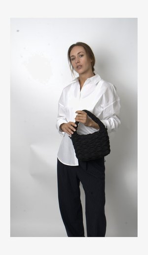 Γυναίκα με λευκό πουκάμισο και μαύρο ραμμένο παντελόνι, κρατώντας μια υφασμάτινη μαύρη τσάντα απέναντι από ένα απλό ανοιχτό φόντο.