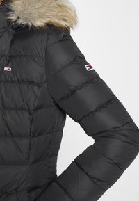 Chaqueta acolchada negra con capucha forrada de piel sintética, textura acolchada y detalles de logo en las mangas. Presenta un diseño ajustado para mayor calidez.