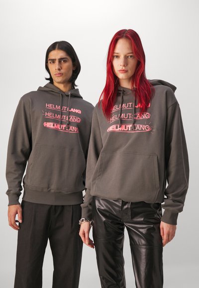 Grå hettegensere med frontlomme og rosa "HELMUT LANG" grafikk, brukt med sorte lærbukser, mot en nøytral bakgrunn.