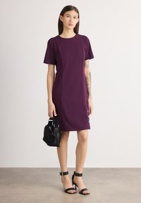 Violeta pieguļošā kleita ar īsām piedurknēm, apaļu dekoltē un sānu pogu detaļām. Modelis tur nelielu melnu kažokādas somiņu un valkā melnus augstpapēžu sandales.