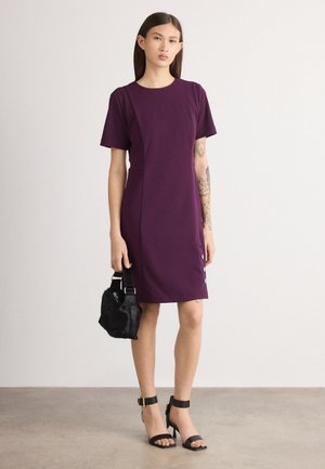 Violetas suknelė, prigludusi prie kūno, su trumpomis rankovėmis, apvaliu iškirpimu ir šoninėmis mygtukų detalėmis. Modelis laiko mažą juodą kailinę rankinę ir dėvi juodus aukštakulnius sandalus.