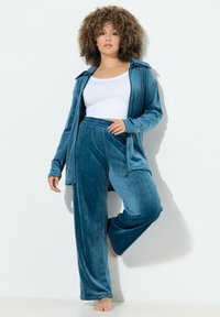 Dvoudílný sametový outfit v barvě smaragdové. Obsahuje bundu se zipem a límcem a kalhoty s elastickým pasem. Měkká textilie, uvolněný střih.