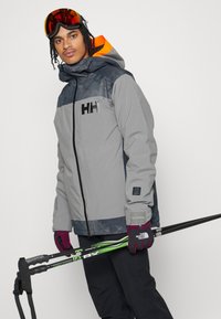 Veste isolée gris et gris foncé avec capuche, arborant un logo audacieux, portée avec un pantalon sombre, des gants violets et des bâtons de ski.