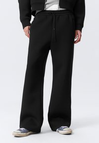 Persona con pantalones de chándal anchos negros con cordón, combinados con una chaqueta negra de cremallera y zapatillas de lona azules desgastadas, sobre fondo blanco.
