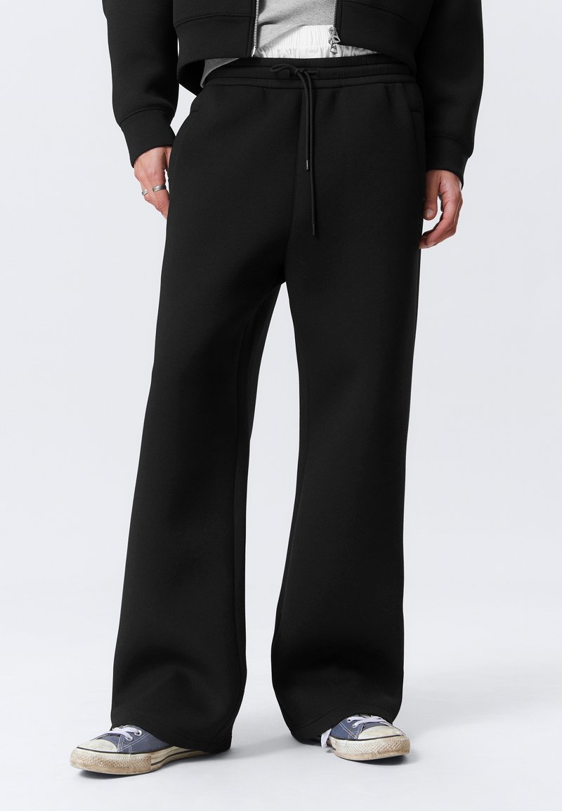 Persona con pantalones de chándal anchos negros con cordón, combinados con una chaqueta negra de cremallera y zapatillas de lona azules desgastadas, sobre fondo blanco.