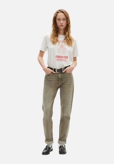 Camisa gráfica blanca con estampado rojo, jeans verde claro, cinturón negro y mocasines de cuero negro. El modelo está de pie con las manos en las caderas.