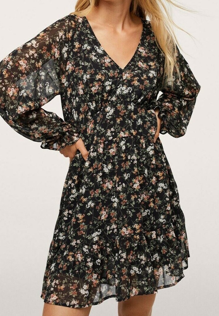 Femme portant une robe noire fleurie avec des manches longues transparentes, un décolleté en V et un ourlet à volants, mains posées sur les hanches.