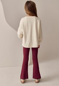 Pulover oversized de culoare crem, cu mâneci lungi, asociat cu pantaloni burgund cu tăietură evazată. Textură moale și design casual, cu detalii vizibile la tiv.