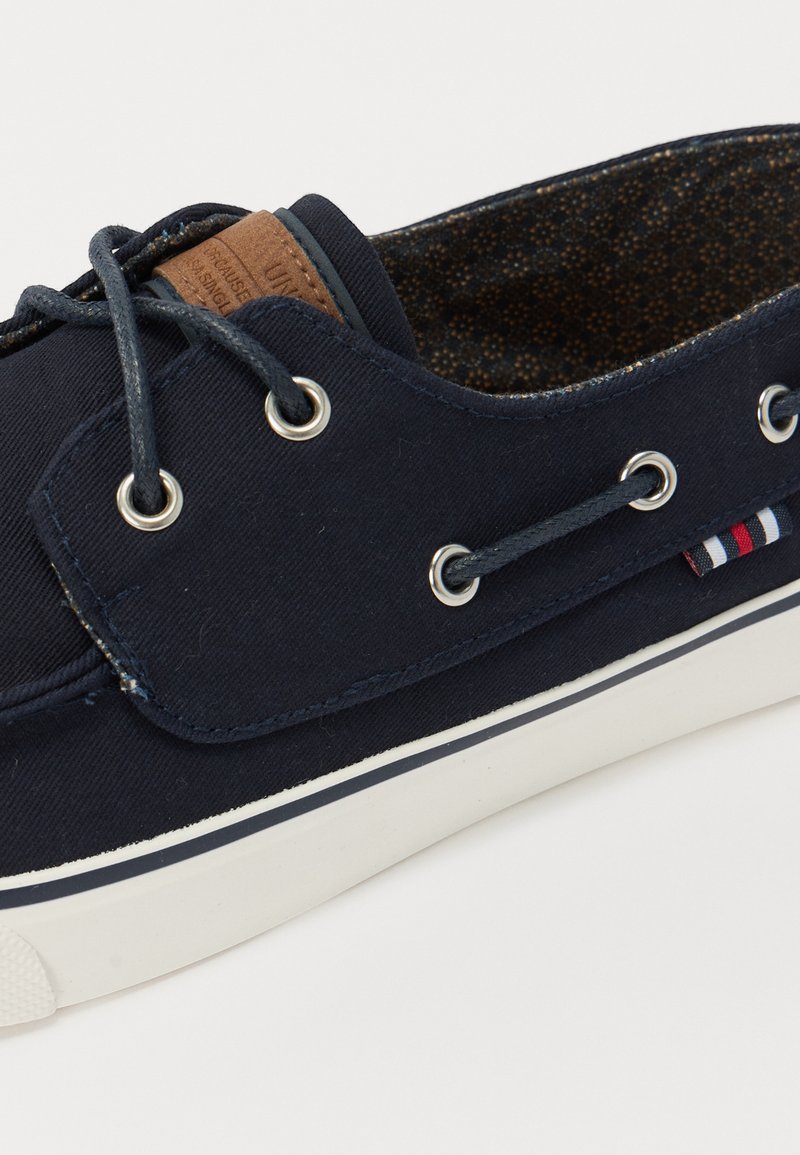 Zapatilla de lona azul marino con ojales metálicos, suela de goma, parche de cuero y detalle a rayas cerca de la suela. Interior texturizado.