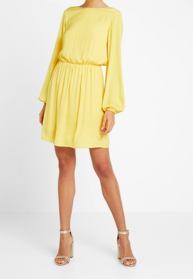 Vestido amarillo con mangas largas abullonadas y cintura fruncida. Hecho de una tela ligera, presenta una falda hasta la rodilla. La modelo lleva tacones dorados.