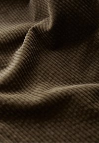 Tessuto in corduroy marrone con una texture a coste prominenti, che crea un aspetto morbido e ondulato. La luce si riflette sulla superficie, esaltando la profondità.