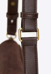 Brązowy skórzany pasek o teksturowanej powierzchni, z metalowym złotym zapięciem i detalami przeszycia, przymocowany do brązowego zamszowego korpusu torby.