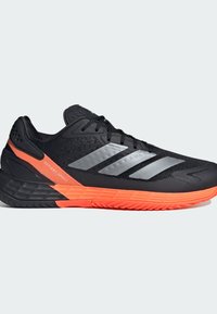 Μαύρο και πορτοκαλί αθλητικό παπούτσι με μαύρες κορδόνες, τρεις ανακλαστικές ασημί λωρίδες στο πλάι και υφή στη σόλα. Το κείμενο "Defiant Speed" κοντά στη φτέρνα.