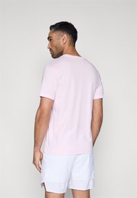 Ανοιχτό ροζ βαμβακερό t-shirt με στρογγυλή λαιμόκοψη, κοντά μανίκια και άνετη γραμμή. Φοράει με λευκά σορτς, εμφανιζόμενο από πίσω.