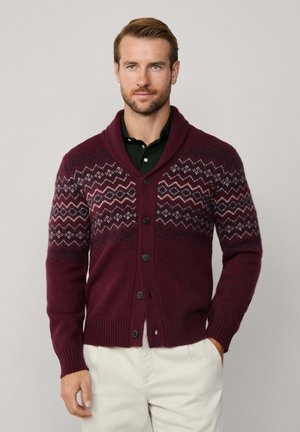 Burgunderfarbener Strickcardigan mit Schalkragen, schwarzen Knöpfen und einem Zickzack-Fair-Isle-Muster in verschiedenen Creme- und Marineblau-Tönen auf der Brust und den Ärmeln.