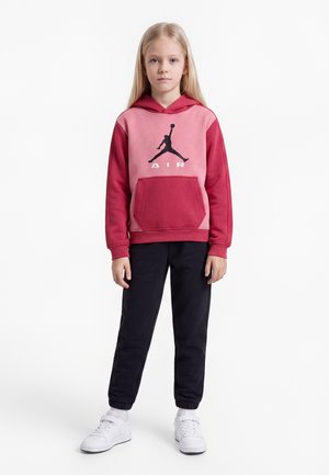 Ξανθό παιδί όρθιο, φοράει ροζ και κόκκινο hoodie με λογότυπο μπάσκετ, μαύρο παντελόνι, λευκές κάλτσες και λευκά αθλητικά παπούτσια σε λευκό φόντο.