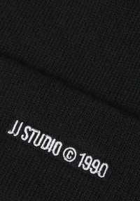 Zwarte gebreide beanie met ribbeltextuur en witte geborduurde merklogo "JJ STUDIO © 1990" nabij de onderrand.