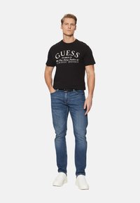 Camiseta de algodón negra con el logo blanco de "GUESS", combinada con unos vaqueros de mezclilla azules. El modelo lleva zapatillas blancas y está de pie frente a un fondo liso.