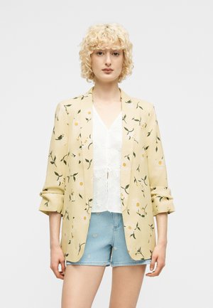 Femme aux cheveux blonds bouclés et courts portant un blazer floral jaune pâle, un chemisier blanc et un short en denim bleu clair avec broderie florale.