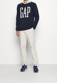 Uomo che indossa un pullover navy blu GAP, una camicia bianca con colletto, pantaloni beige, calzini bianchi e sneaker navy blu, in piedi contro uno sfondo bianco.