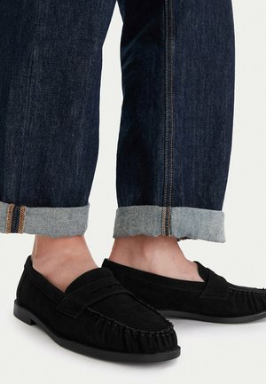 Osoba nosząca ciemnoniebieskie, podwinięte dżinsy oraz czarne mokasyny zamszowe, stojąca na jasnym tle.