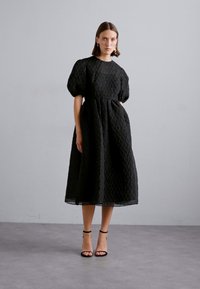 Black textured dress with a diamond pattern, short puff sleeves, and a knee-length A-line silhouette. Worn with black strappy sandals.