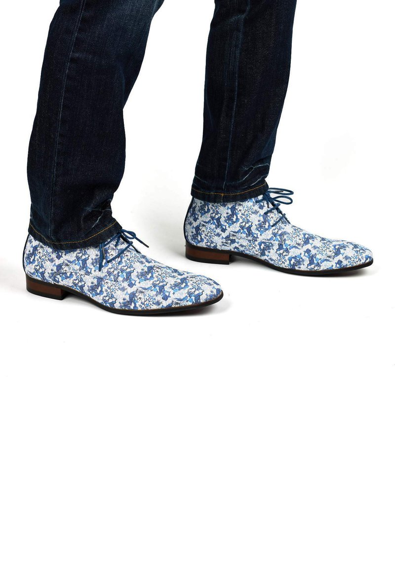 Blue floral-patterned shoes made of fabric with a smooth texture, featuring a pointed toe and thin laces, paired with dark denim jeans.