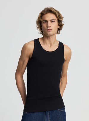 Młody mężczyzna o kręconych włosach, ubrany w czarny, bezrękawnik z prążkowanej dzianiny i niebieskie dżinsy, stoi na tle jasnego, jednolitego tła.