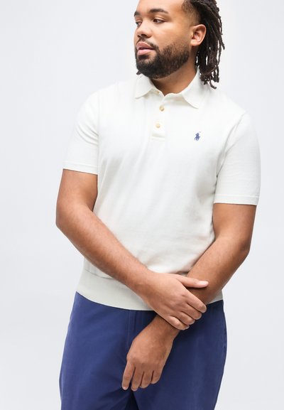 Άνδρας με γενειάδα και στα dreadlocks, φοράει λευκό πόλο με κοντά μανίκια και μπλε παντελόνι, στέκεται με τα χέρια σταυρωμένα μπροστά σε απλό φόντο.