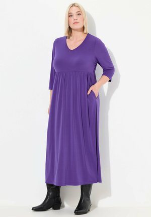 Robe violette, longueur genoux, en tissu doux, avec un décolleté en V, des manches trois-quarts, et des poches latérales. Portée avec des bottes noires.