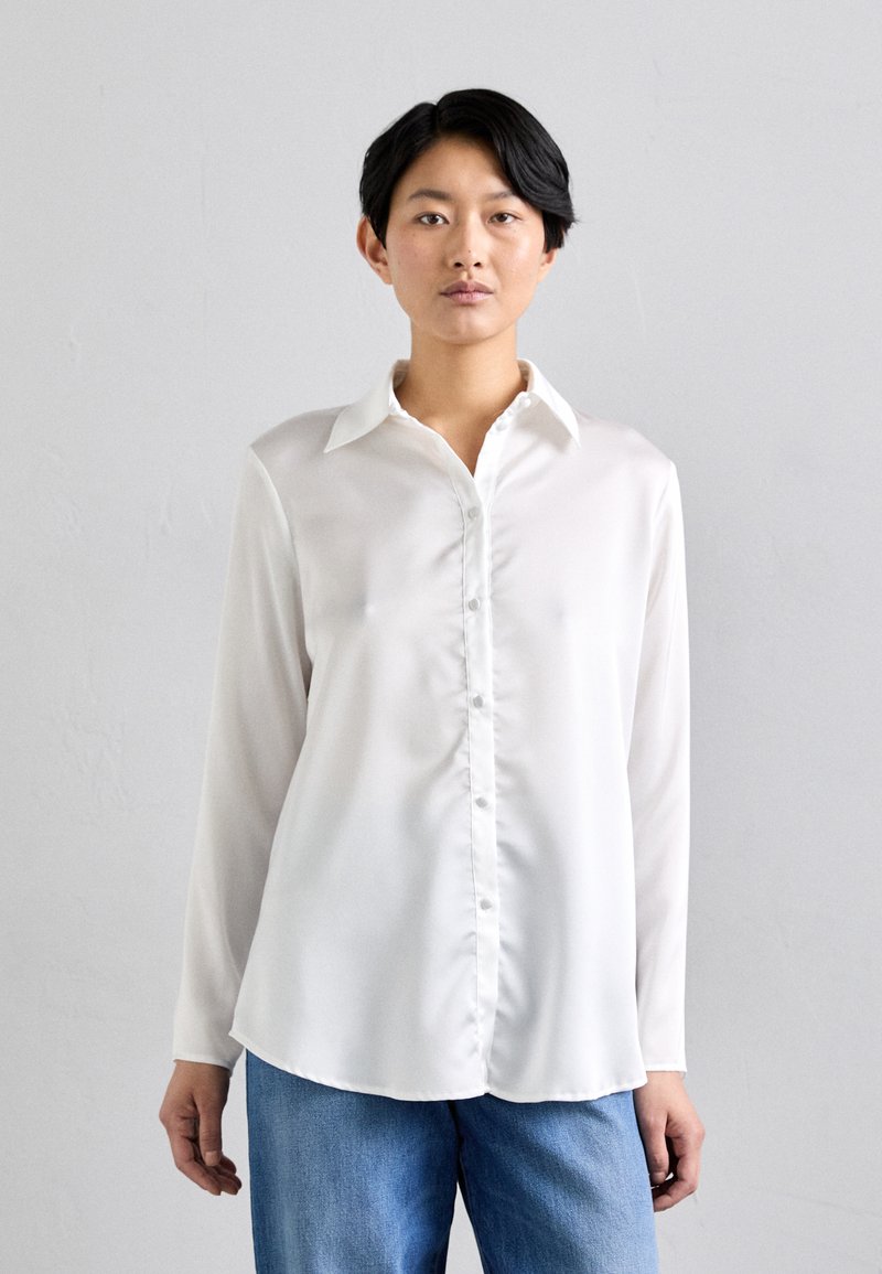 Camicia bianca a maniche lunghe, realizzata in tessuto morbido, presenta un colletto classico, bottoni frontali e un orlo curvo, abbinata a un paio di pantaloni in denim blu.