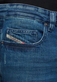 Siniset farkut, joissa on takatasku, jossa on näkyvä etiketti, niittidetaljit ja haalistunut, teksturoitu pinta.