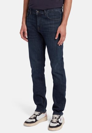 Jean bleu foncé avec une coupe slim, présentant un design classique à cinq poches et une fermeture à bouton métallique. Porté avec des baskets noires et blanches.