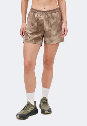 Personne portant un short marron tie-dye, des chaussettes blanches montantes et des baskets Adidas vert olive, debout devant un fond clair uni.