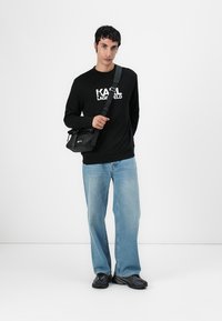 Schwarzer Sweatshirt mit weißem "KARL LAGERFELD"-Logo, blaue Denim-Jeans und schwarze Umhängetasche. Das Model steht vor einem neutralen Hintergrund.
