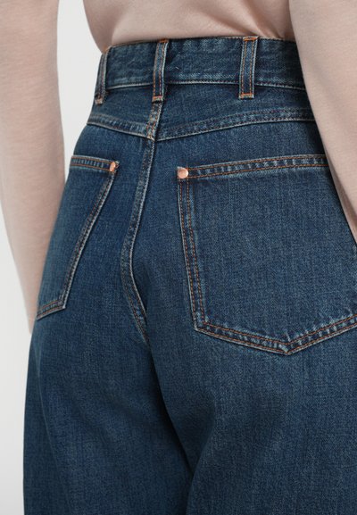 Jeans de mezclilla azul oscuro de tiro alto con costuras naranjas contrastantes, dos bolsillos traseros y remaches de cobre visibles. Textura suave.