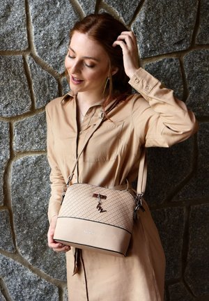 Woman with reddish hair wearing a beige dress, holding a beige quilted crossbody bag with a charm, standing against a stone wall background.
