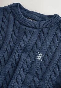 Merekas sinise kaabelkootud kampsun, millel on tekstuuriga muster, sooniline krae ja eest valge tikitud logo.