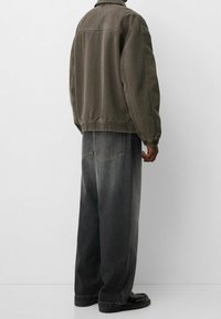 Přiléhavá olivově zelená bunda s žebrovaným lemem a manžetami, nošena přes volné tmavě šedé džíny. Hladká tkanina s viditelnými detaily šití.