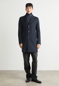 Casaco em mistura de lã azul-marinho com um design elegante, de botão único na frente, lapelas em bico e dois bolsos frontais. Usado sobre uma jaqueta interna com fecho éclair.