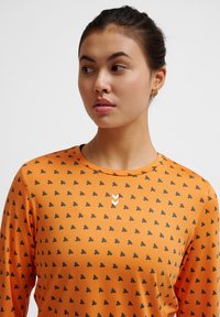 Oranje longsleeve shirt met een herhalend zwart patroon van kleine vormen, met een wit logo op de borst. Zacht textuur, ronde halslijn.