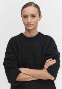 Svart, fluffig tröja med rundad halslinning och 3/4-långa ärmar. Modellen står med armarna i kors, mot en neutral bakgrund.