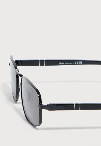 Μαύρα ορθογώνια γυαλιά ηλίου με λεπτά μεταλλικά πλαίσια και σκουρόχρωμους φακούς, εμφανιζόμενα από πλάγια γωνία σε λευκό φόντο.