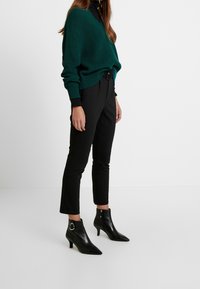 Zielony sweter z prążkowanego dzianiny, czarne dopasowane spodnie, oraz czarne szpilki za kostkę z metalowymi akcentami w kolorze srebrnym.