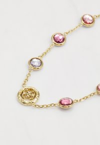 Gouden ketting met ovale, roze en paarse facette stenen. Voorzien van een decoratieve ronde sluiting met een bloemenmotief en een schakelkettinkje.